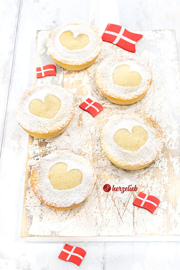 5 kleine gefüllte Kuchen nach einem Cremelinser Rezept aus Dänemark. Alle Kuchen sind mit Puderzucker bestäubt, dabei wurde jeweils ein Herz ausgespart. Zwischen den Kuchen liebe kleine Dannebrog. Angerichtet auf einem weißen Holzbrett