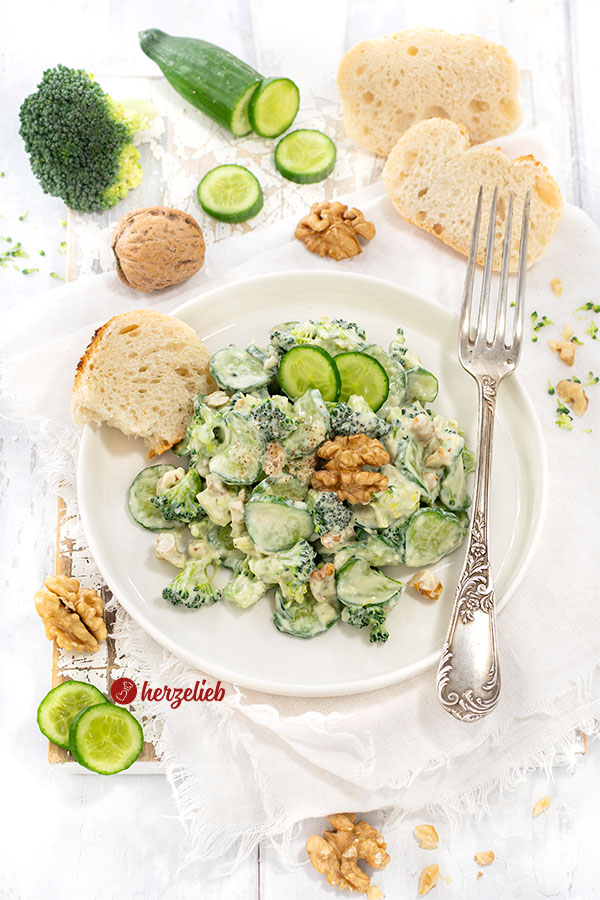 Ein Teller mit einem grünen Salat mit einem cremigen Dressing nach einem Gurken-Brokkoli-Salat von herzellieb. Angerichtet mit Brot, dekoriert mit Gurke und Walnuss. Auf dem Tellerrand eine Silbergabel.