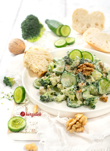 Ein weißer Teller mit einem grünen Salat mit einem cremigen Dressing nach einem Gurken-Brokkoli-Salat von herzellieb. Serviert mit Brot, dekoriert mit einer Walnüssen und Gurke.