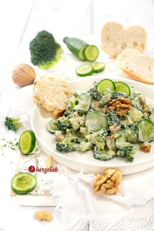 Ein weißer Teller mit einem grünen Salat mit einem cremigen Dressing nach einem Gurken-Brokkoli-Salat von herzellieb. Serviert mit Brot, dekoriert mit einer Walnüssen und Gurke.