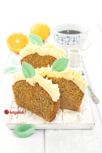 3 Scheiben Karottenkuchen mit Frostinghaube nach einem Orangen-Karotten-Kastenkuchen Rezept von herzelieb auf einem weißen Holzbrett. Dekoriert mit grünen Blättern aus Esspapier. Im hintergrund Orangen, eine Tasse Kaffee und ein Milchkännchen. Seitlich eine Kuchengabel