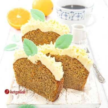 3 Scheiben Karottenkuchen mit Frostinghaube nach einem Orangen-Karotten-Kastenkuchen Rezept von herzelieb auf einem weißen Holzbrett. Dekoriert mit grünen Blättern aus Esspapier. Im hintergrund Orangen, eine Tasse Kaffee und ein Milchkännchen. Seitlich eine Kuchengabel