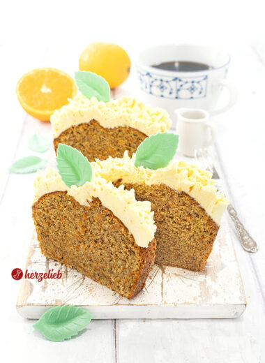 3 Scheiben Karottenkuchen mit Frostinghaube nach einem Orangen-Karotten-Kastenkuchen Rezept von herzelieb auf einem weißen Holzbrett. Dekoriert mit grünen Blättern aus Esspapier. Im hintergrund Orangen, eine Tasse Kaffee und ein Milchkännchen. Seitlich eine Kuchengabel