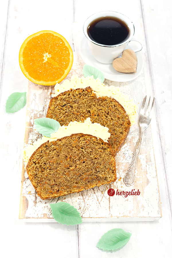 2 Scheiben saftiger Karottenkuchen mit Frostinghaube nach einem Orangen-Karotten-Kastenkuchen Rezept von herzelieb liegen schräg übereinander auf einem weißen Holzbrett. Seitlich davon eine Kuchengabel. Dekoriert mit grünen Blättern aus Esspapier. Im Hintergrund eine halbe Orange und, eine Tasse Kaffee.