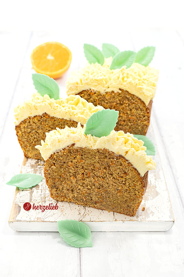 2 dicke Scheiben Karottenkuchen mit Frostinghaube nach einem Orangen-Karotten-Kastenkuchen Rezept von herzelieb stehen aufrecht vor dem geschnittenen Kuchen auf einem weißen Holzbrett. Seitlich davon eine Kuchengabel. Dekoriert mit grünen Blättern aus Esspapier. Im Hintergrund eine halbe Orange.