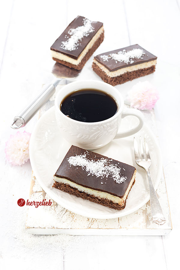 Ein Kuchenstück nach einem Schoko-Kokos-Kuchen von herzelieb. Angerichtet mit einer Tasse Kaffe auf einem ovalen Teller. Auf dem Tellerrand eine Kuchengabel. Im Hintergrund 2 weitere Stücke Kuchen. 