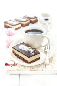 1 Stück Kuchen mit einer Tasse Kaffee nach einem Schoko-Kokos-Kuchen Rezept von herzelieb. Im Hintergrund eine Platte mit drei weiteren Kuchentücken