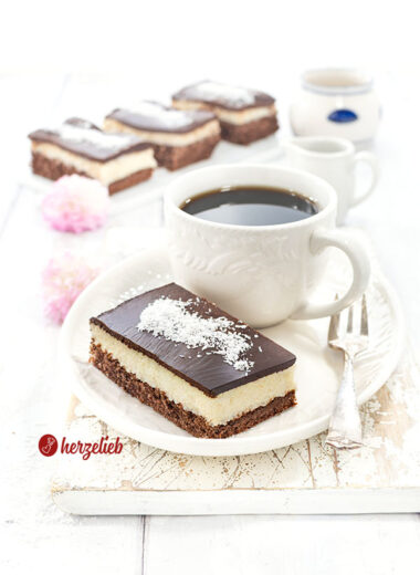 1 Stück Kuchen mit einer Tasse Kaffee nach einem Schoko-Kokos-Kuchen Rezept von herzelieb. Im Hintergrund eine Platte mit drei weiteren Kuchentücken