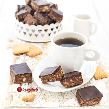 Eine Kleine Tasse Espresso auf einem kleinen, weißen Porzellanteller. Auf dem Untertellr liegen zwei Schokohappen mit Schokoladenguss. Davon und dahinter lieben noch zwei Stücke davon. Im Hintergrund eine Schale mit den knusprigen Schokowürfeln