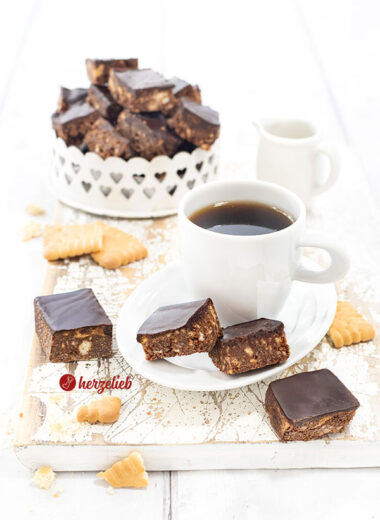 Eine Kleine Tasse Espresso auf einem kleinen, weißen Porzellanteller. Auf dem Untertellr liegen zwei Schokohappen mit Schokoladenguss. Davon und dahinter lieben noch zwei Stücke davon. Im Hintergrund eine Schale mit den knusprigen Schokowürfeln