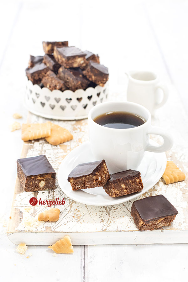 Eine Kleine Tasse Espresso auf einem kleinen, weißen Porzellanteller. Auf dem Untertellr liegen zwei Schokohappen mit Schokoladenguss. Davon und dahinter lieben noch zwei Stücke davon. Im Hintergrund eine Schale mit den knusprigen Schokowürfeln