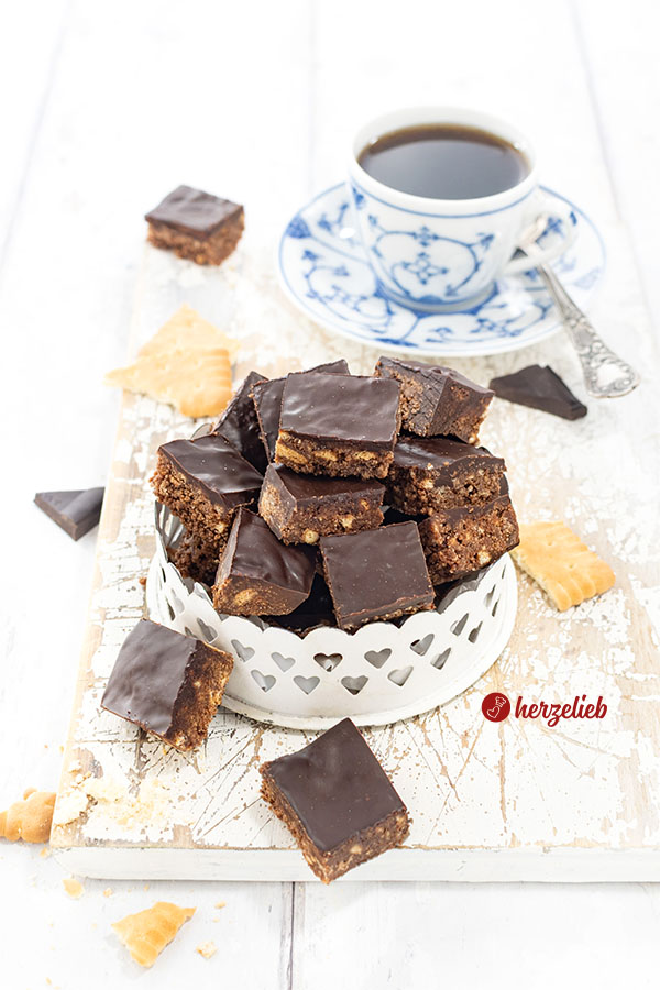 Eine runde Metallschale mit Herzen, in die kleine, knusprige Schokohappen mit Schokoladenguss geschichtet sind. Im Hintergrund eine Tasse mit Kaffee.
