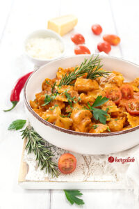 Am rechten Bildrand ist eine Schale mit einer Tomaten-Hähnchen-Pasta. Dekoriert mit roter Chili, Tomaten, Parmesan und Rosmarinzweigen. Bestreut mit gehackter Petersilie.