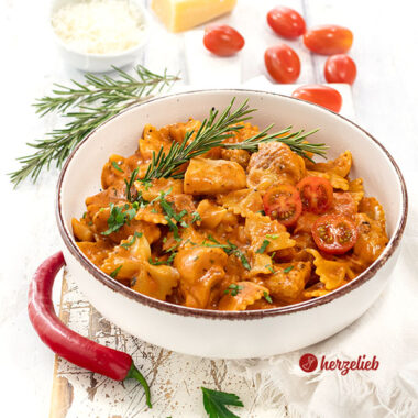 Leckeres One-Pot-Gericht nach einem Tomaten-Hähnchen Pasta Rezept von herzelieb, angerichtet in ener Schale. Dekoriert mit Chili, Rosmarin, Tomaten, Petersilie. Im Hintergrund Parmesan.