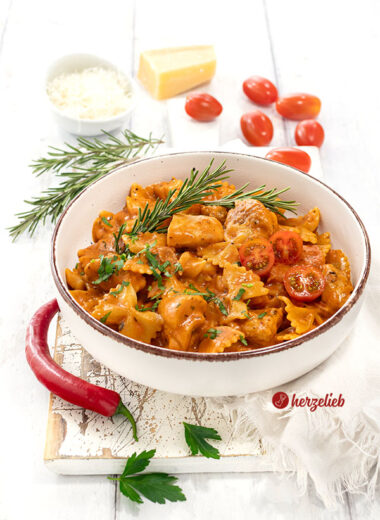 Leckeres One-Pot-Gericht nach einem Tomaten-Hähnchen Pasta Rezept von herzelieb, angerichtet in ener Schale. Dekoriert mit Chili, Rosmarin, Tomaten, Petersilie. Im Hintergrund Parmesan.