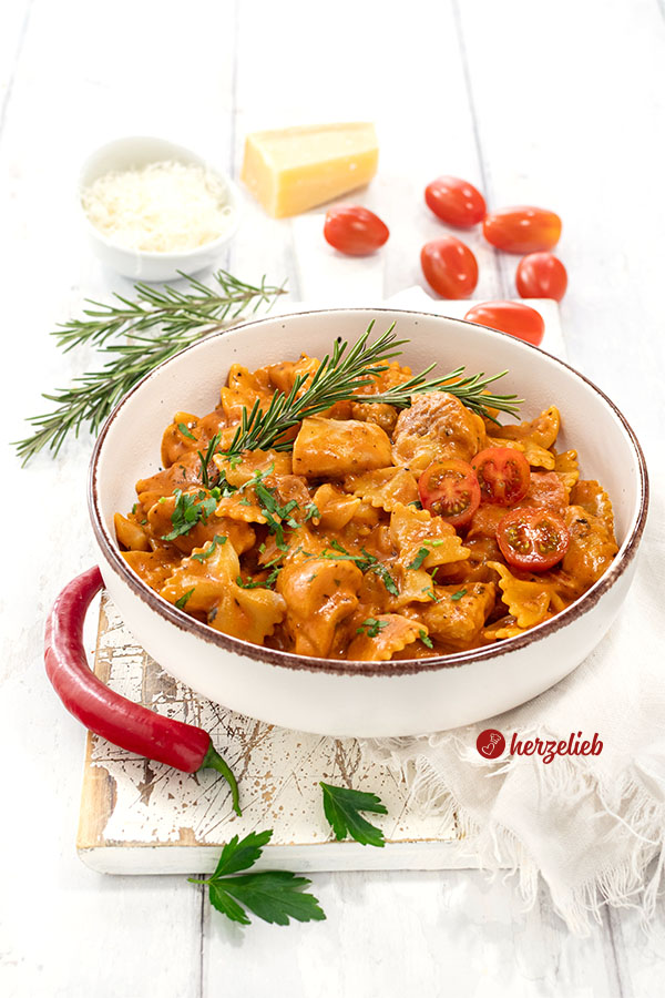 Leckeres One-Pot-Gericht nach einem Tomaten-Hähnchen Pasta Rezept von herzelieb, angerichtet in ener Schale. Dekoriert mit Chili, Rosmarin, Tomaten, Petersilie. Im Hintergrund Parmesan.