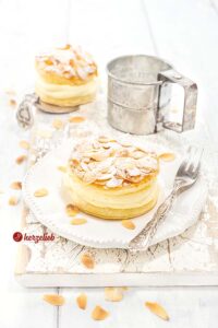 Ein Bienenstichtörtchen aus Blätterteig mit Mandeln und Puderzucker auf einem weißen Teller. Auf dem Tellerrand eine Gabel, im Hintergrund ein Puderzuckersieb und ein weiteres Törtchen