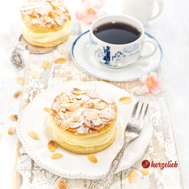 Ein gedeckter Kaffeetisch Im Fokus ein Bienenstichtörtchen aus Blätterteig mit Mandeln obendrauf von herzelieb auf einem Teller. Auf dem Tellerand eine Gbel