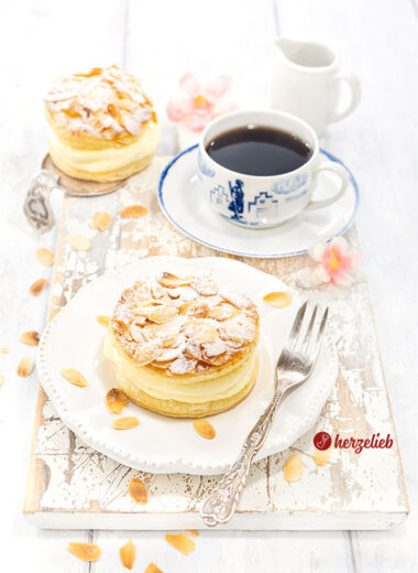 Ein gedeckter Kaffeetisch Im Fokus ein Bienenstichtörtchen aus Blätterteig mit Mandeln obendrauf von herzelieb auf einem Teller. Auf dem Tellerand eine Gbel