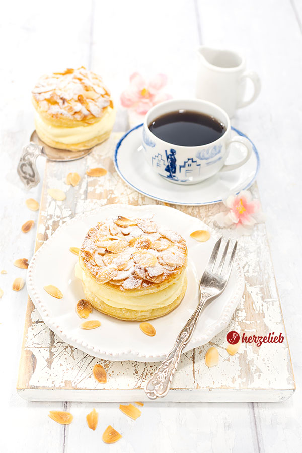 Ein gedeckter Kaffeetisch Im Fokus ein Bienenstichtörtchen aus Blätterteig mit Mandeln obendrauf von herzelieb auf einem Teller. Auf dem Tellerand eine Gabel