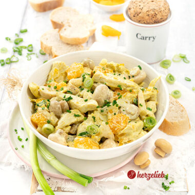 Schale mit Geflügelsalat mit Mandarinen auf einem Teller. Im Hintergrund Brot, ein Currygefäß. Dekoriert mit Lauchzwiebeln, Pilzen und Mandarinen.