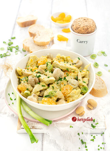 Schale mit Geflügelsalat mit Mandarinen auf einem Teller. Im Hintergrund Brot, ein Currygefäß. Dekoriert mit Lauchzwiebeln, Pilzen und Mandarinen.