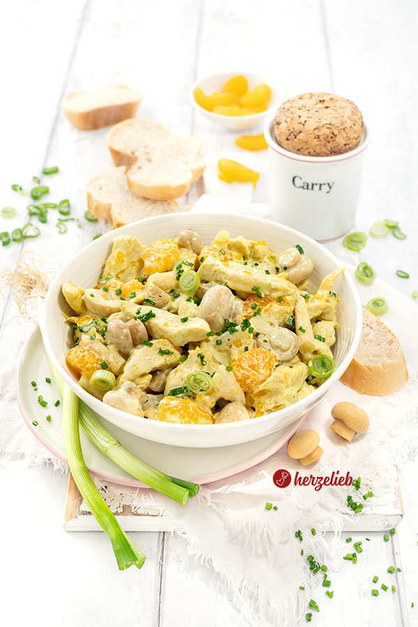 Schale mit Geflügelsalat mit Mandarinen auf einem Teller. Im Hintergrund Brot, ein Currygefäß. Dekoriert mit Lauchzwiebeln, Pilzen und Mandarinen.