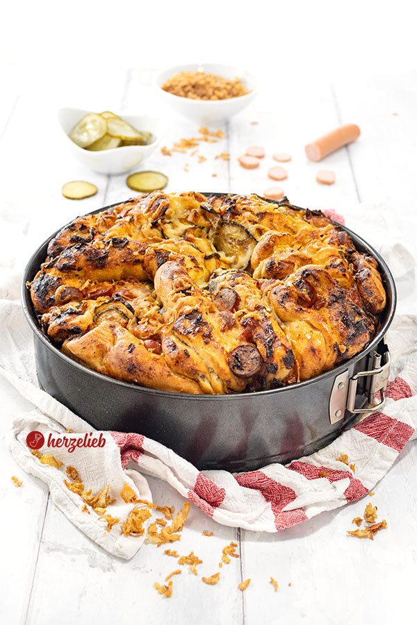 Hotdog-Faltenbrot in einer Springform. Goldbraun gebacken. Angerichtet mit Röstzwiebeln, Gurken und Würstchen
