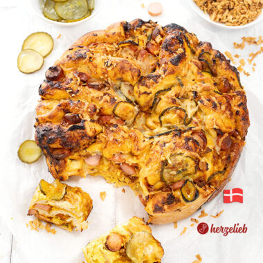 Goldbraun gebackenes Hotdog-Faltenbrot auf weißem Backpapier. Mit Würstchen, Agurker und Röstzwiebeln angerichtet.