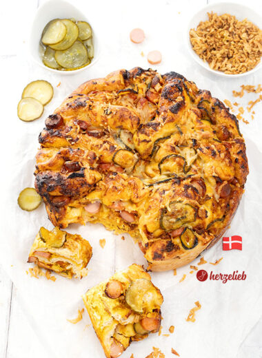 Goldbraun gebackenes Hotdog-Faltenbrot auf weißem Backpapier. Mit Würstchen, Agurker und Röstzwiebeln angerichtet.