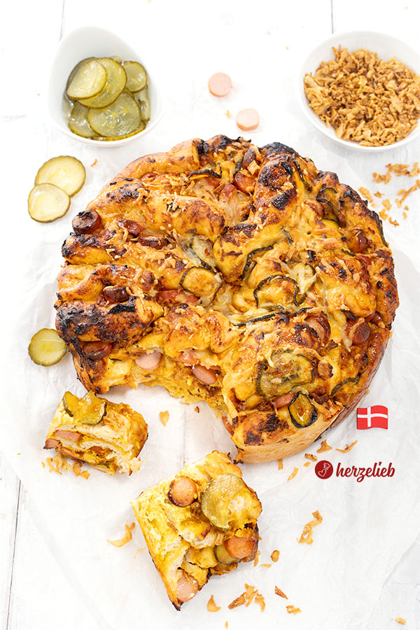 Goldbraun gebackenes Hotdog-Faltenbrot auf weißem Backpapier. Mit Würstchen, Agurker und Röstzwiebeln angerichtet. 