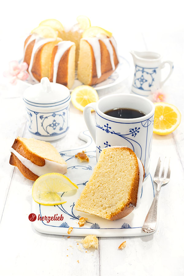 Gedeckter Kaffeetisch, ein Stück Joghurt-Zitronen-Gugelhupf mit Kaffee auf einem Brett. Im Hintergrund der Zitronenkuchen.