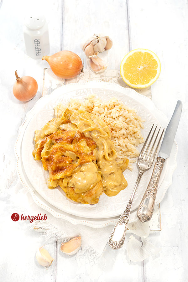 Zwiebel-Sahne-Hähnchen in sahniger Soße auf einem weißen Teller. Besteck auf dem Tellerrand. im Hintergrund Zwiebeln, Knoblauch, eine halbe Zitrone, ein Pfefferstreuer. Von oben fotografiert.