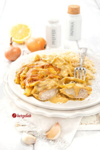 Leckeres Zwiebel-Sahne-Hähnchen mit Reis auf einem weißen Teller. Im Vordergrund ein angeschnittenes Stück Hähnchen, ein Stück auf einer Gabel. Im Hintergrund Zwiebeln, eine halbe Zitrone, eine Knoblauchknolle, zwei Gewürzgefäße.