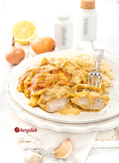 Leckeres Zwiebel-Sahne-Hähnchen mit Reis auf einem weißen Teller. Im Vordergrund ein angeschnittenes Stück Hähnchen, ein Stück auf einer Gabel. Im Hintergrund Zwiebeln, eine halbe Zitrone, eine Knoblauchknolle, zwei Gewürzgefäße.