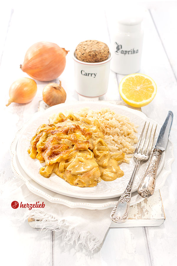 Leckeres Zwiebel-Sahne-Hähnchen mit Zwiebelringen in sahniger Soße. Auf dem Tellerrand Besteck. Im Hintergrund zwei Gewürzgefäße, eine halbe Zitrone, Zwiebeln. Vorn zwei Knoblauchzehen. 