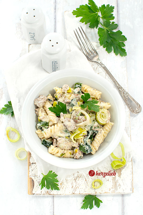 Cremige Hack-Lauch-Nudelpfanne mit Käse auf einem weißen Teller von oben. Dekoriert mit glatter Petersilie. Oben im Hintergrund eine Silbergabel. 