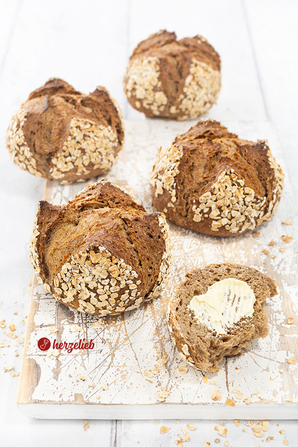 4 Havregrynsboller aus Dänemark auf einem Holzbrett. Mit Haferflocken bestreut. Vorn ein Stück vom Haferflockenbrötchen mit Butter. 