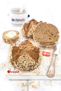 Vorn ein Havregrynsboller oder Haferflockenbrötchen aus Dänemark. Knackig gebacken, dunkle Krume. Seitlich ein Messer und ein Dannebrog. Hinten Schmalztopf.
