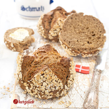 Vorn ein Havregrynsboller oder Haferflockenbrötchen aus Dänemark. Knackig gebacken, dunkle Krume. Seitlich ein Messer und ein Dannebrog. Hinten Schmalztopf.