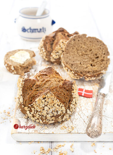 Vorn ein Havregrynsboller oder Haferflockenbrötchen aus Dänemark. Knackig gebacken, dunkle Krume. Seitlich ein Messer und ein Dannebrog. Hinten Schmalztopf.