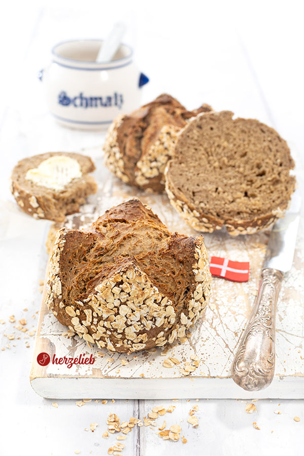 Vorn ein Havregrynsboller oder Haferflockenbrötchen aus Dänemark. Knackig gebacken, dunkle Krume. Seitlich ein Messer und ein Dannebrog. Hinten Schmalztopf.