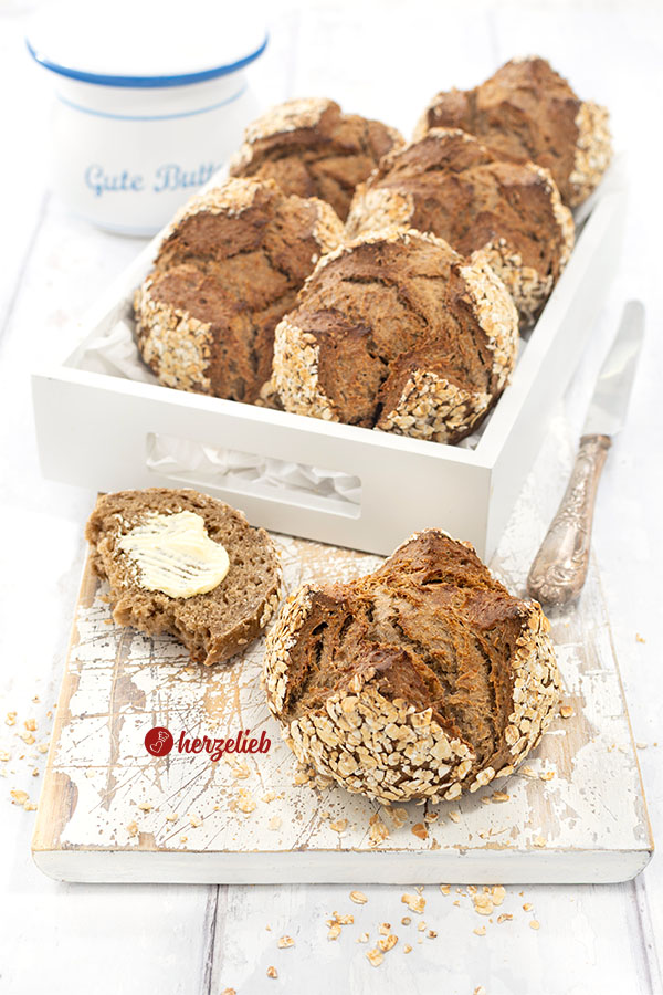 Vorn ein Havregrynsboller oder Haferflockenbrötchen aus Dänemark. Knackig gebacken, dunkle Krume. Seitlich ein Messer und ein Dannebrog. Hinten Schmalztopf.