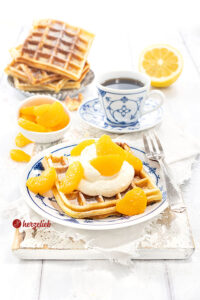 Käsekuchenwaffeln auf einem gedecktem Kaffetisch. Im Vordergrund eine Waffel mit Sahne und Mandarinen auf einem blau-weißen Teller.