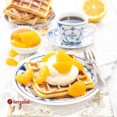 Käsekuchenwaffeln auf einem gedecktem Kaffetisch. Im Vordergrund eine Waffel mit Sahne und Mandarinen auf einem blau-weißen Teller.