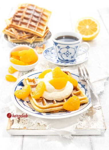 Käsekuchenwaffeln auf einem gedecktem Kaffetisch. Im Vordergrund eine Waffel mit Sahne und Mandarinen auf einem blau-weißen Teller.