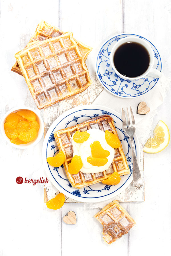 Kaffeetisch mit blauem Geschirr von oben. Eine der Käsekuchenwaffeln in der Mitte mit Sahne und Mandarinen. Hinten links weitere Waffeln.