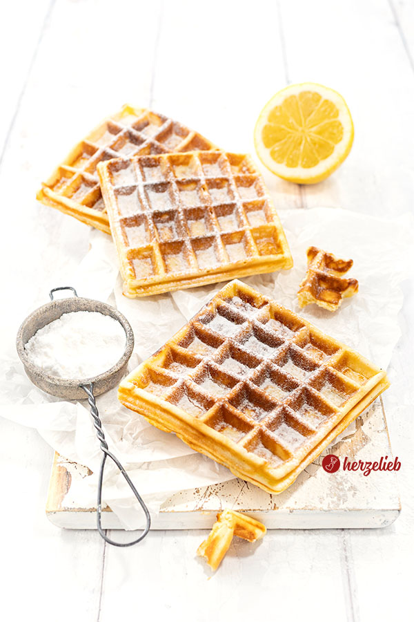 Drei Käsekuchenwaffeln auf einem weißen Holzbrett, dass mit zerknülltem Papier belegt ist. Vorne links ein Sieb mit Puderzucker, hinten eine Zitrone