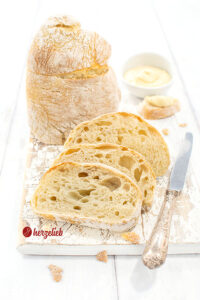 Drei Scheiben Pantoffelbrot vor einem auf der Schnittfläche stehenden Brotlaib. Seitlich neben dem Ciabatta ein Silbermesser, hinten ein Dip.