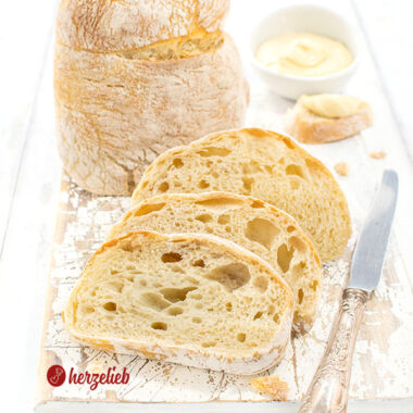 Drei Scheiben Pantoffelbrot vor einem auf der Schnittfläche stehenden Brotlaib. Seitlich neben dem Ciabatta ein Silbermesser, hinten ein Dip.
